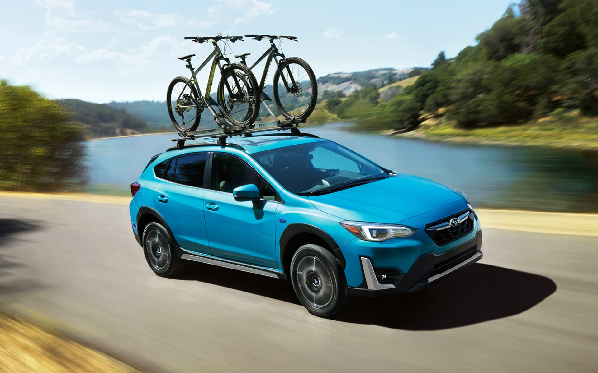 A blue Crosstrek Hybrid with two bicycles on its roof rack driving beside a river | Dyer Subaru in Vero Beach FL